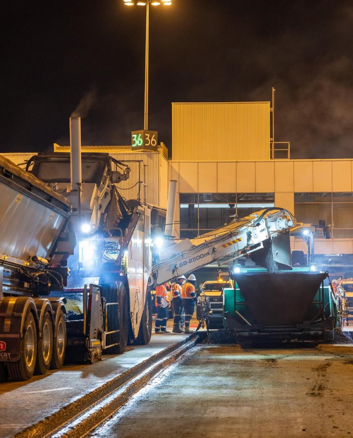 Asphalt delivery and paving equipment working under lights near airport terminal buildings